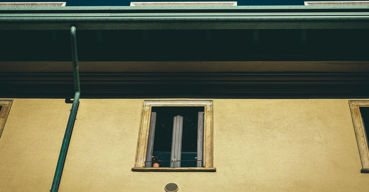 Upward view of a yellow stucco building facade with a narrow open window, dark roof overhang, and green drainpipe.