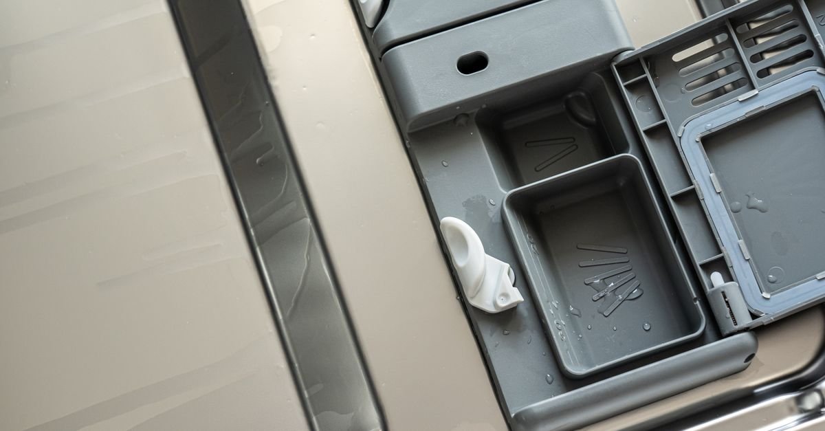 Open gray plastic dishwasher detergent dispenser with water droplets on its compartments.