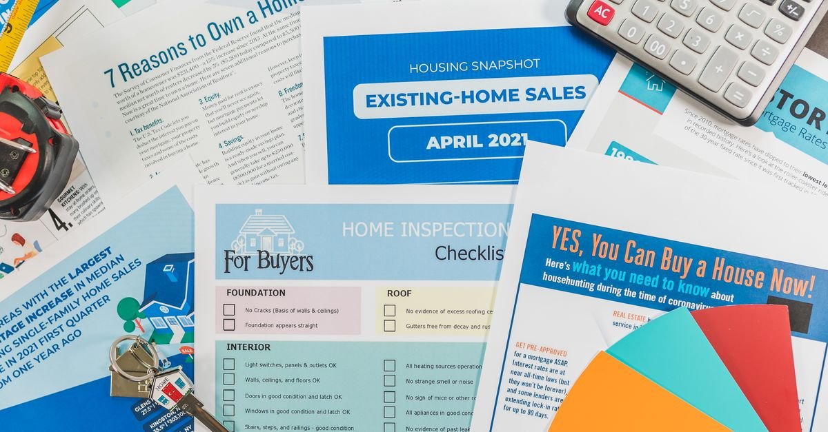 Overlapping home-buying documents, a home inspection checklist, a housing snapshot, a calculator, keys, and colorful folders are arranged on a tabletop.