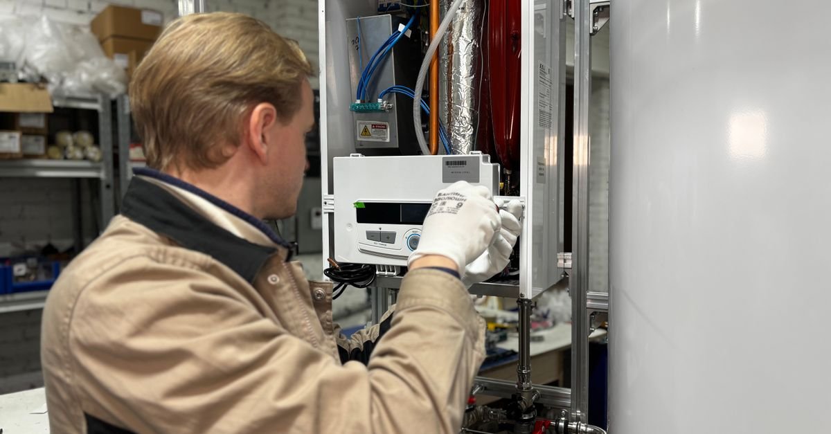 A technician wearing gloves works on the control panel of an open heating unit in a workshop.