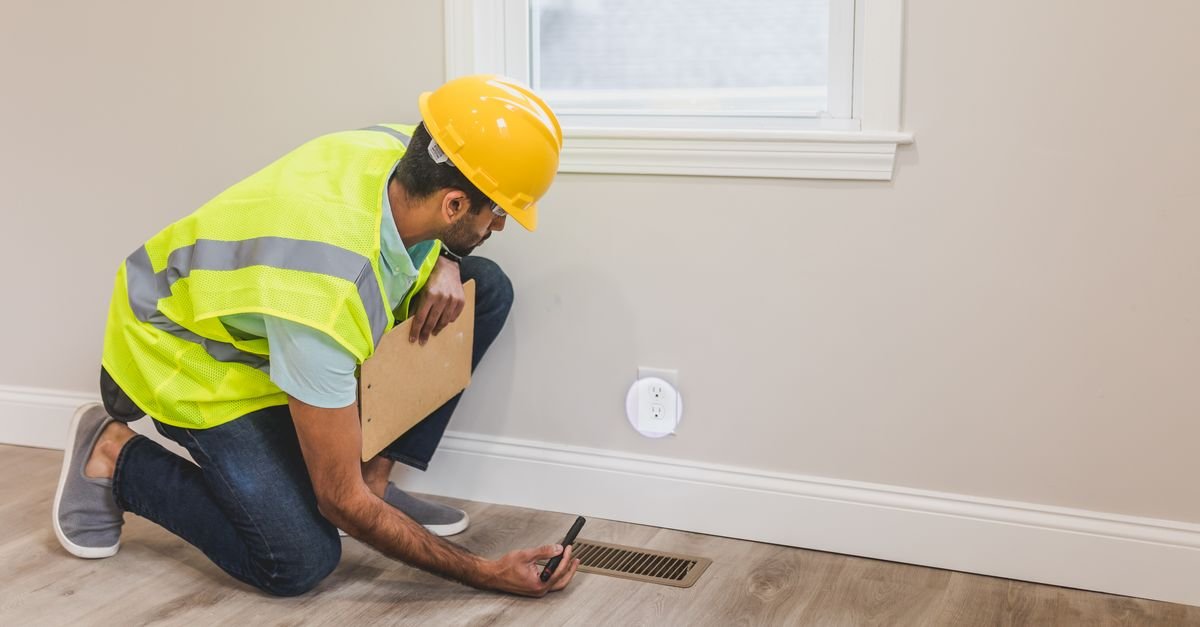 A worker in a yellow hard hat and reflective vest kneels on a wooden floor beside a floor vent, holding a tool.