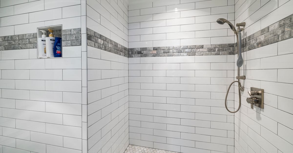 A tiled shower enclosure with white subway tiles, gray accent strips, a recessed shelf holding bottles, and a wall-mounted shower system.