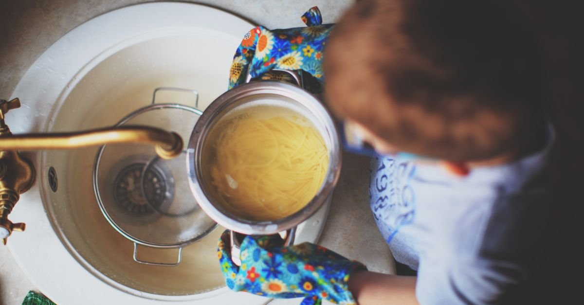 Top-down view of a person wearing floral oven mitts pouring cooked spaghetti from a pot into a colander in a kitchen sink.