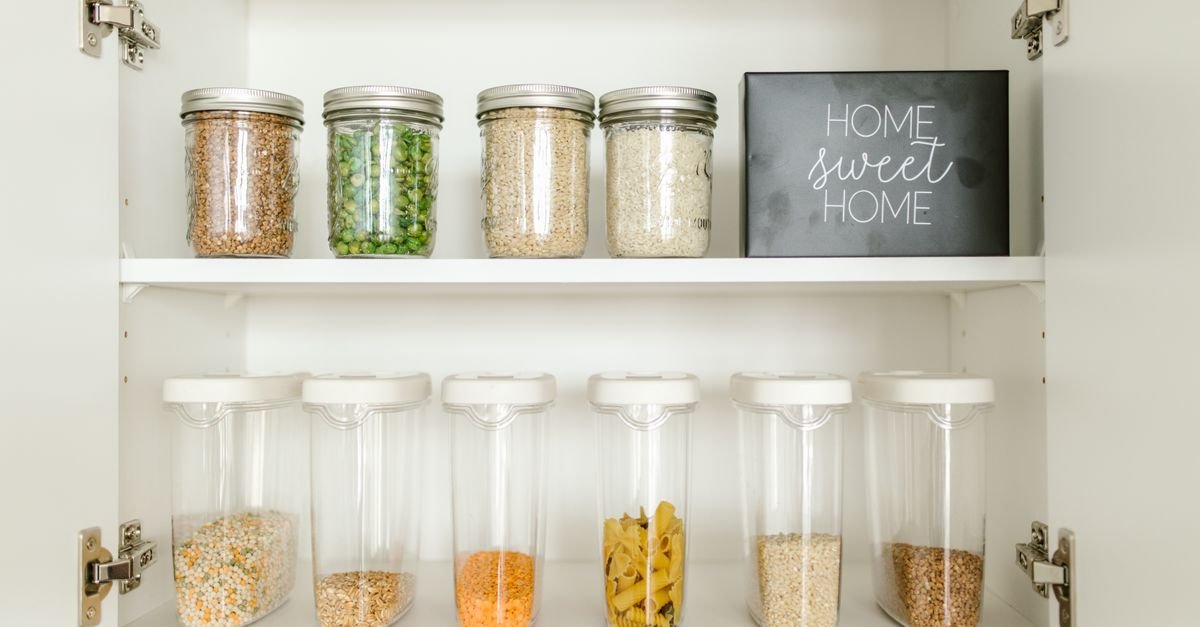 Open white pantry cabinet with shelves of glass jars containing dry foods and a black sign reading “Home Sweet Home.”