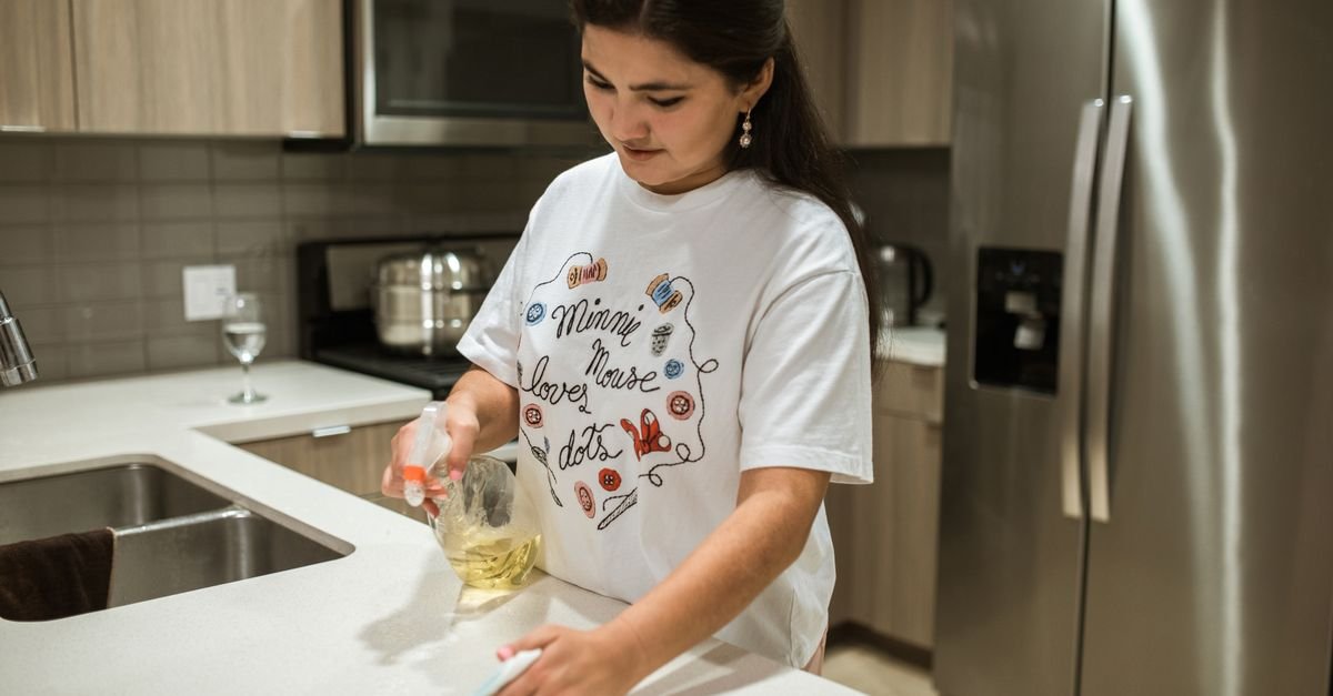 A woman in a white graphic t-shirt wipes down a white kitchen counter with a spray bottle and cloth in a modern kitchen with stainless steel appliances.
