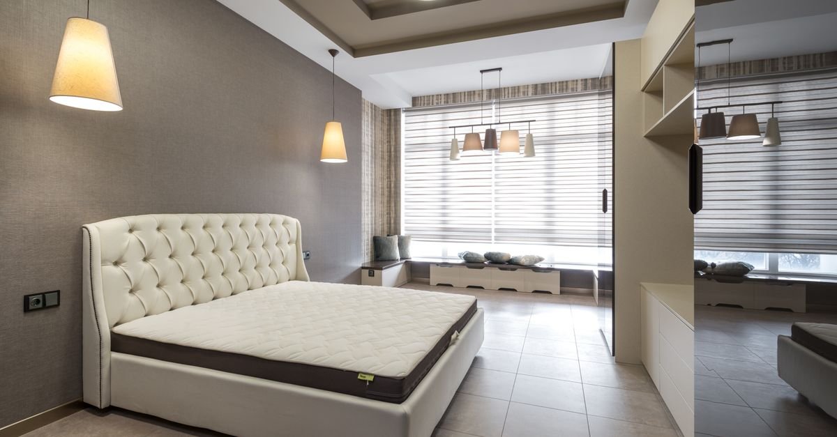 Modern bedroom with a tufted cream bed, pendant lights, large windows with blinds, built-in storage, and a mirrored wall.