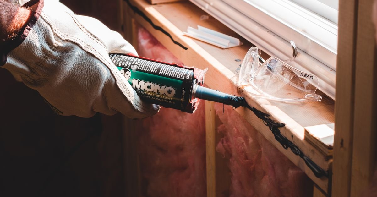 A gloved hand applies black sealant along a wooden window sill beside exposed insulation.
