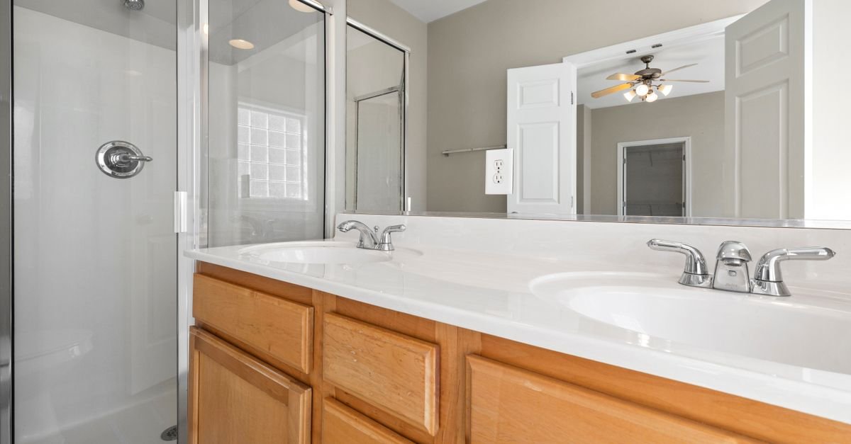 A bright bathroom features a glass-enclosed shower, a double-sink vanity with wooden cabinets, and a large wall mirror.