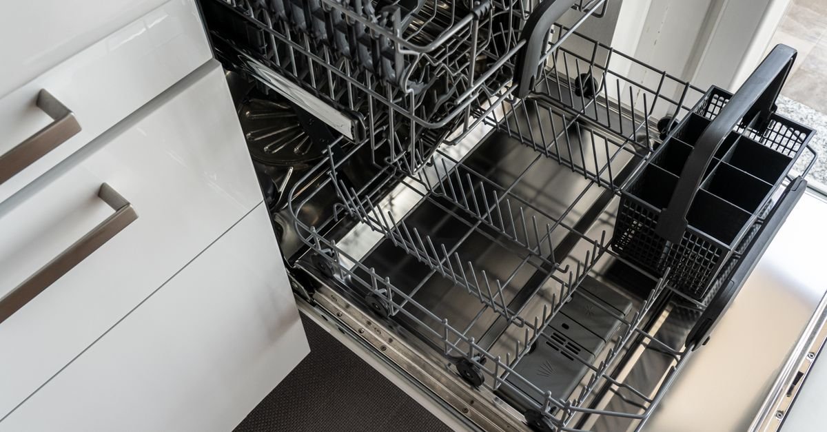 An open dishwasher with empty racks and a utensil basket beside white kitchen cabinets.