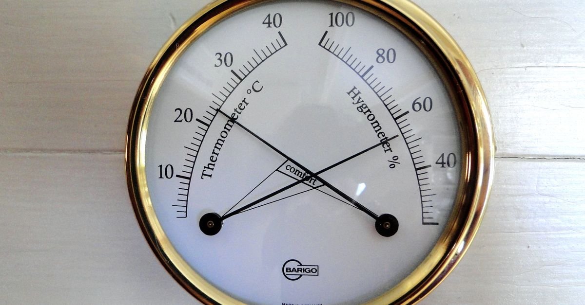 A round analog thermometer and hygrometer gauge with black needles, white face, and gold rim mounted on a light wooden surface.